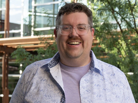 Image of Dr. Rob Groves, a tall white man wearing glasses, smiling, and wearing a lavendar t-shirt with a blue and white patterned button down.