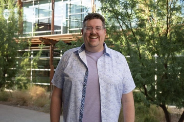 Image of Dr. Rob Groves, a tall white man wearing glasses, smiling, and wearing a lavendar t-shirt with a blue and white patterned button down.