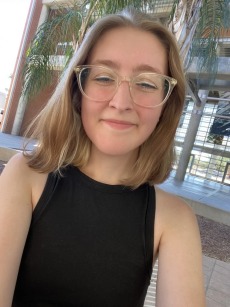 Photo of Lyssa. They are wearing a black shirt, clear glasses, and have shoulder length blonde hair. 