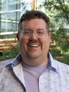 Image of Dr. Rob Groves, a tall white man wearing glasses, smiling, and wearing a lavendar t-shirt with a blue and white patterned button down.
